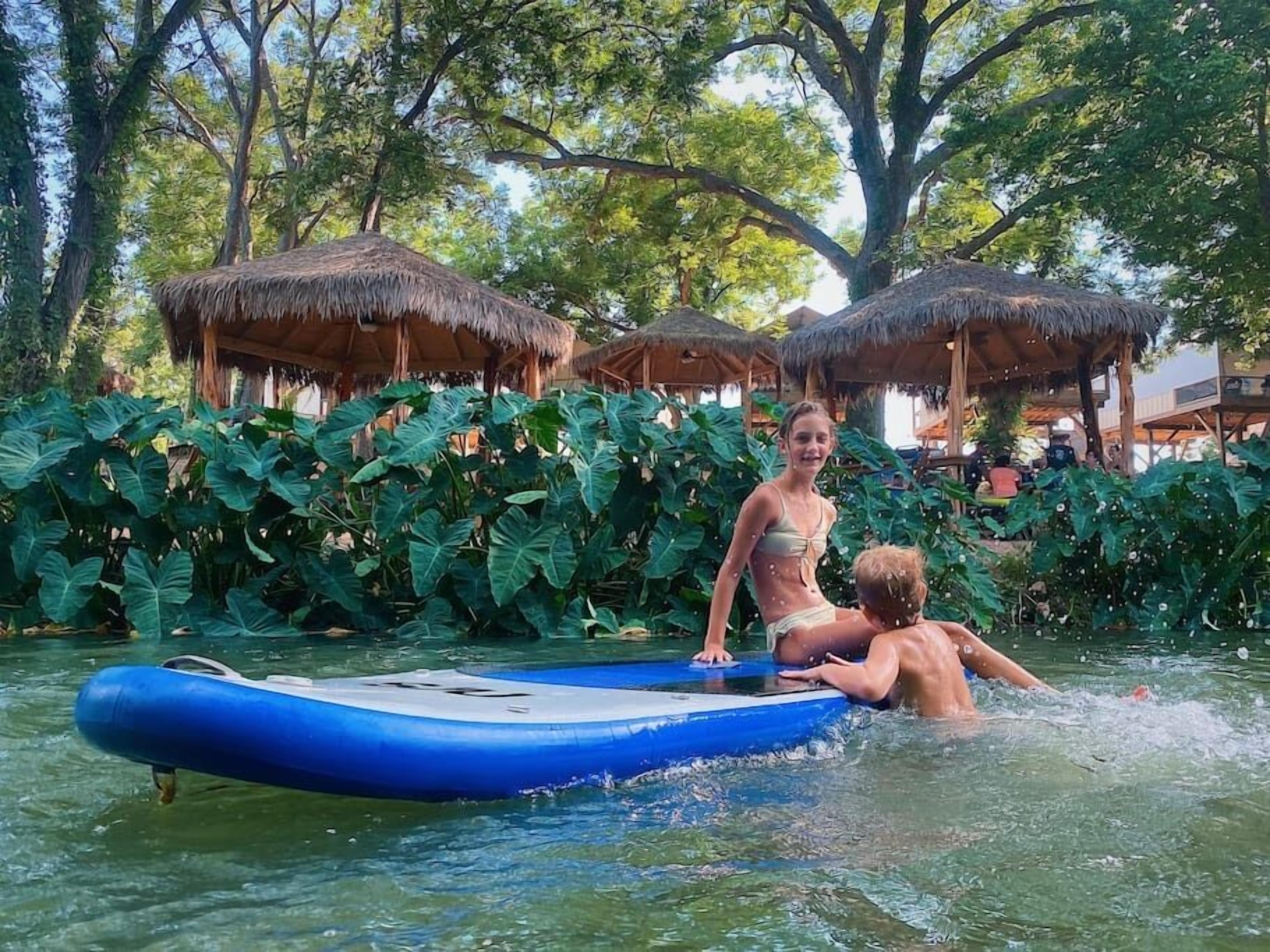 Un espace piscine agrémenté d’un radeau gonflable bleu, entouré d’une végétation tropicale et de constructions à toits de chaume.