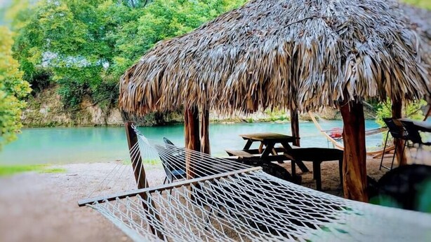 A thatched-roof hut with a hammock and picnic table by a calm body of water, surrounded by lush greenery. - #50 Son's River Ranch Safari Cabin (Guadalupe County)