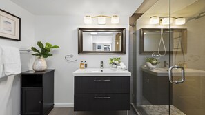 A modern bathroom with a white sink, dark cabinets, a large mirror, and a glass shower enclosure. - Condo with Oceanfront Pool & Direct Beach Access at the Roney Palace (Miami Beach)