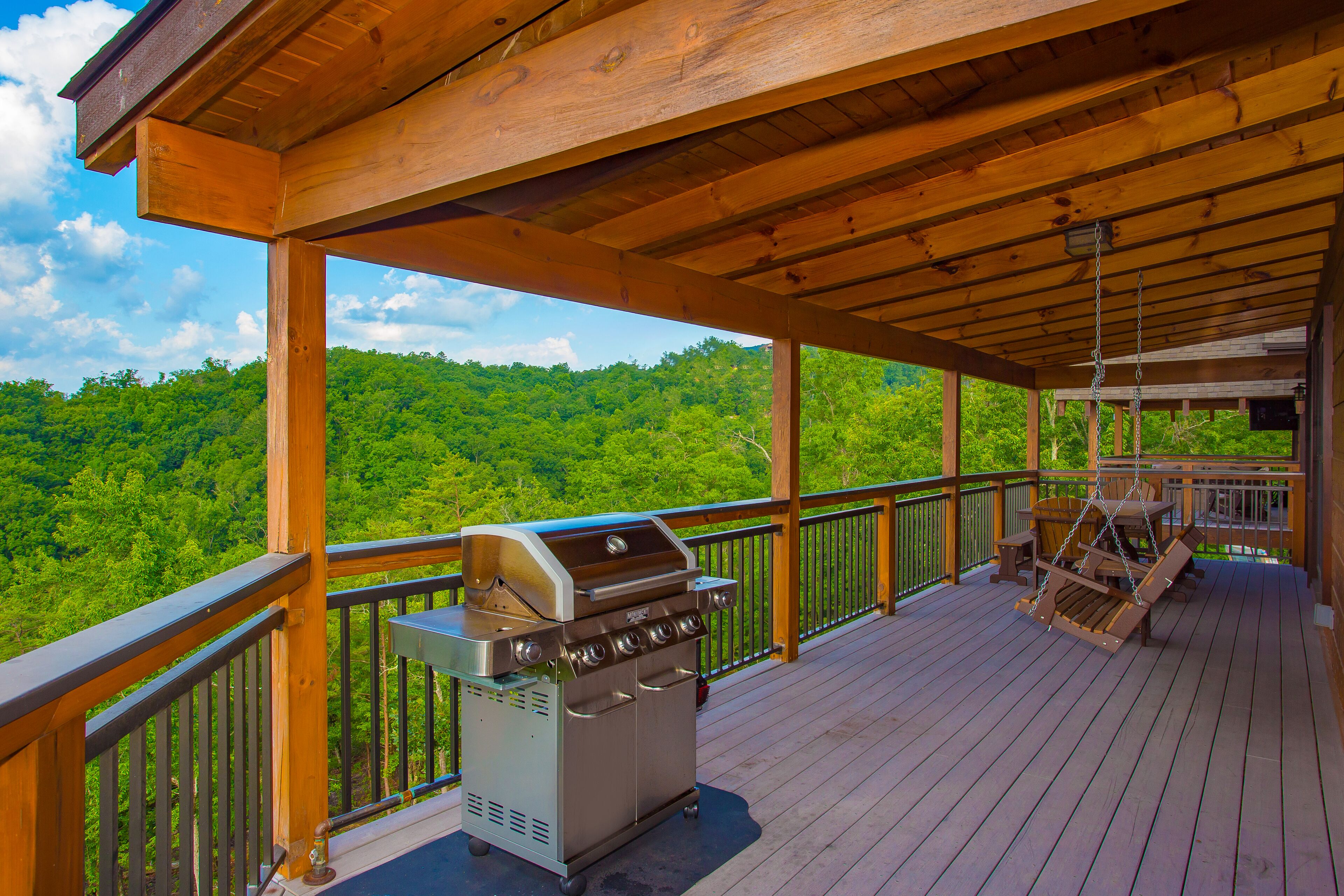 A covered deck with a stainless steel grill, wooden railings, and a view of a forested hillside.