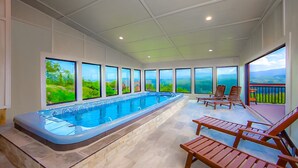 Une piscine avec vue sur un paysage verdoyant, grâce à de grandes fenêtres, des chaises longues en bois et un sol carrelé.