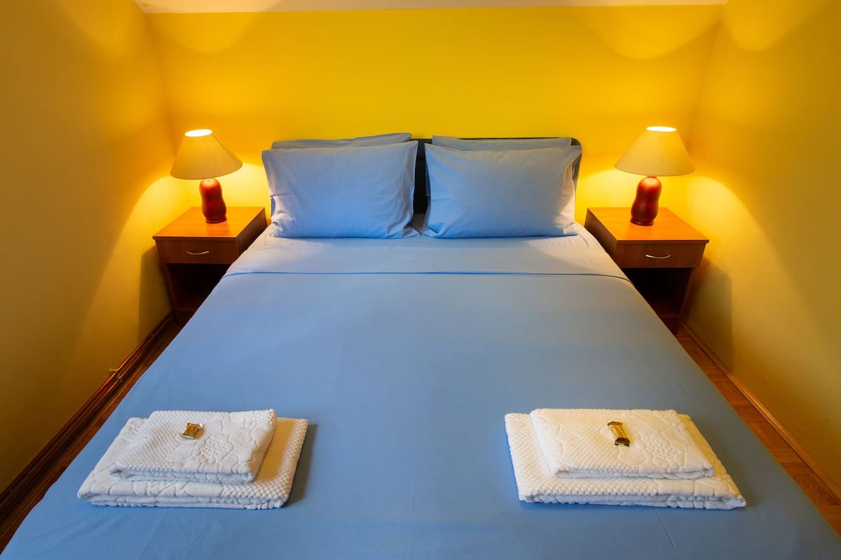 A neatly made bed with blue bedding, two pillows, and two folded towels with a key on top. There are two bedside lamps on wooden nightstands.
