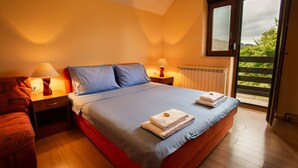 Un dormitorio con una cama grande, dos mesitas de noche con lámparas, una silla roja y vistas a la vegetación a través de una puerta corredera.