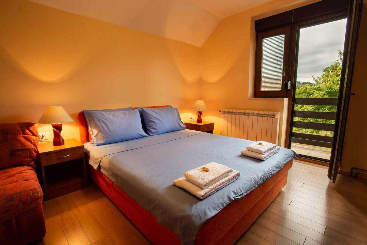 A bedroom with a large bed, two bedside tables with lamps, a red chair, and a view of greenery through a sliding door.