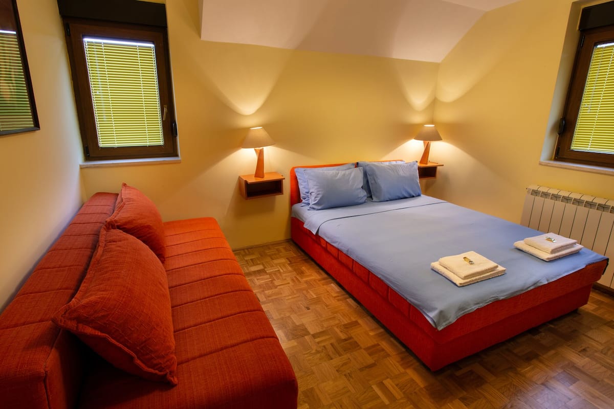 A bedroom with a bed, two bedside tables with lamps, a red sofa, and a window with blinds.
