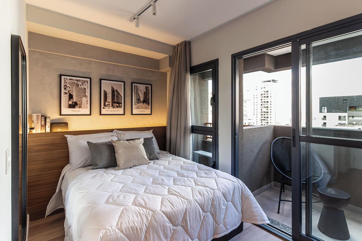 A modern bedroom with a large bed, a balcony with a chair, and cityscape views.