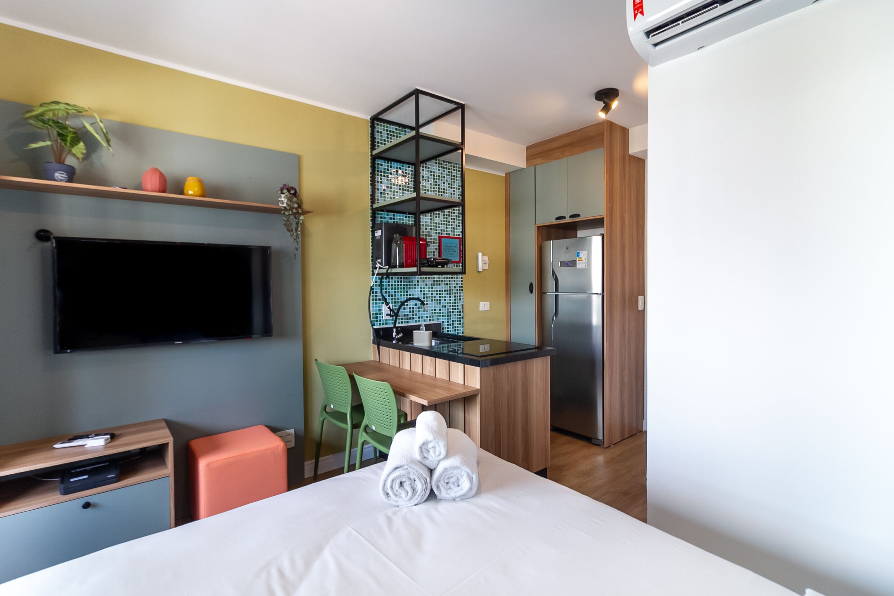 A hotel room with a bed, a TV, a desk, a chair, a small kitchen area, and a shelf with decorative items.