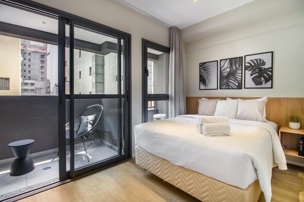 A modern hotel room with a large bed, a glass door leading to a balcony, and a view of the cityscape.