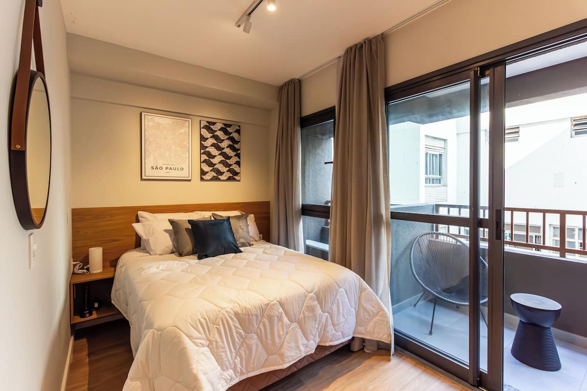 A hotel room with a large bed, a bedside table with a lamp, a framed map of São Paulo, and a balcony with a chair and table.