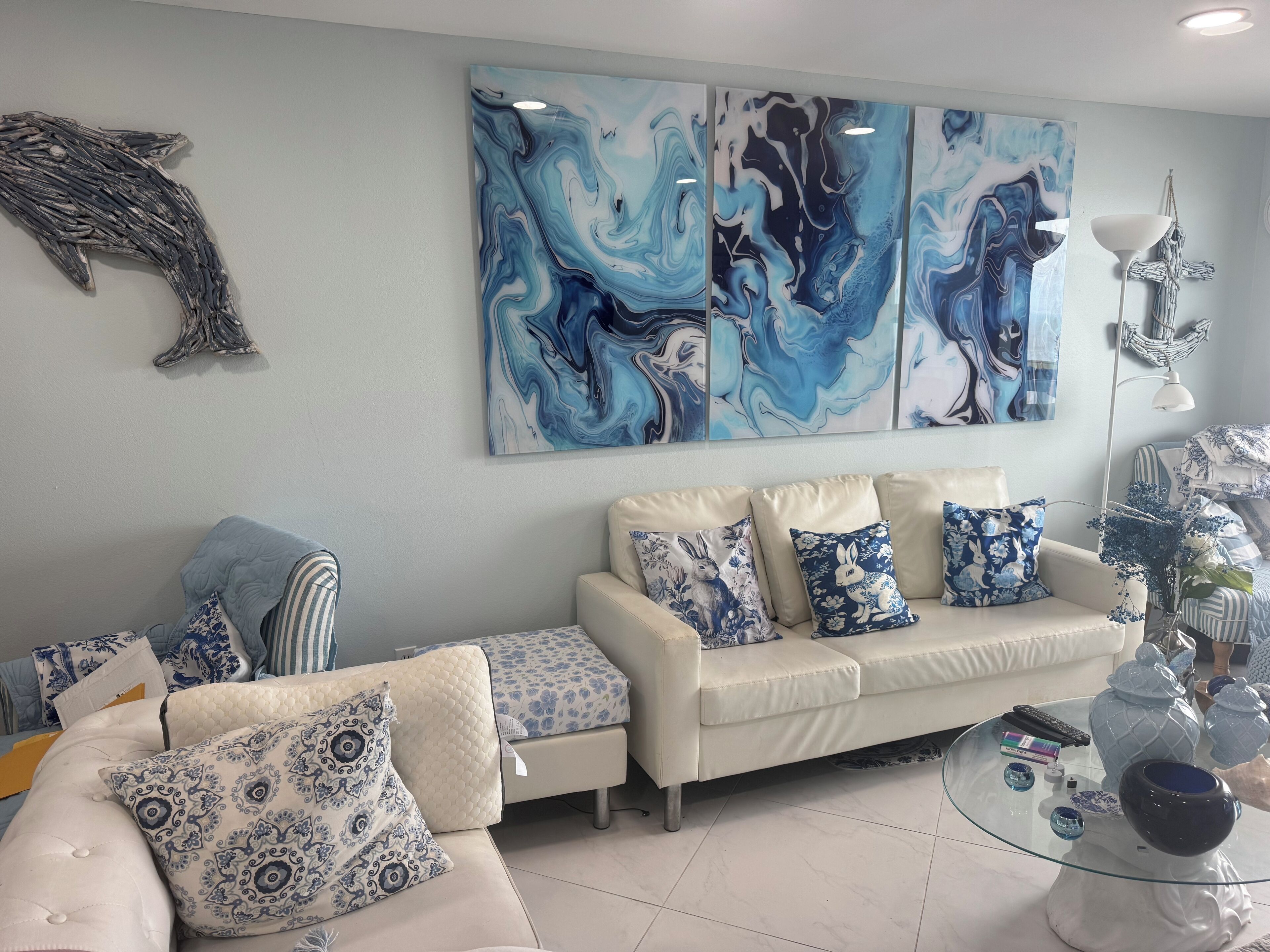 A living room with a white sofa, blue and white patterned cushions, a glass coffee table with decorative items, and abstract blue and white wall art.