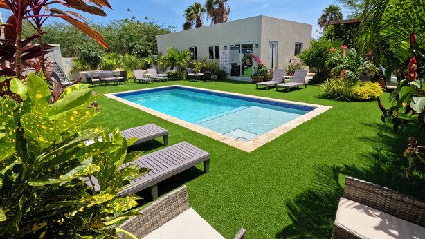 A modern outdoor pool area with artificial grass, lounge chairs, and a small building in the background.