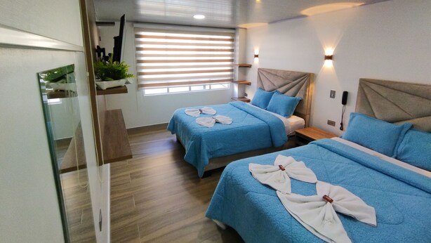 Een hotelkamer met twee bedden, elk met blauw beddengoed en witte handdoeken. Er is een raam met zonwering, een nachtkastje met een lamp, en een spiegel aan de muur.