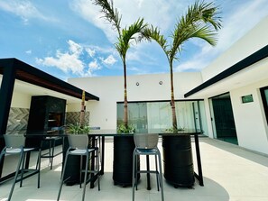 Uma área moderna para refeições ao ar livre, com mesa e cadeiras de vidro, cercada por palmeiras e um edifício branco com amplas janelas.