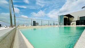 A rooftop pool with clear turquoise water, surrounded by a modern building with large glass windows and a view of the city skyline. - COMODO MONOAMBIENTE EN EQUIPETROL CON PISCINA EN CONDOMINIO! (Santa Cruz de la Sierra)