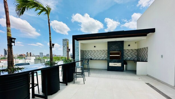 A modern outdoor kitchen area with a built-in oven, seating, and a view of the city skyline. - COMODO MONOAMBIENTE EN EQUIPETROL CON PISCINA EN CONDOMINIO! (Santa Cruz de la Sierra)