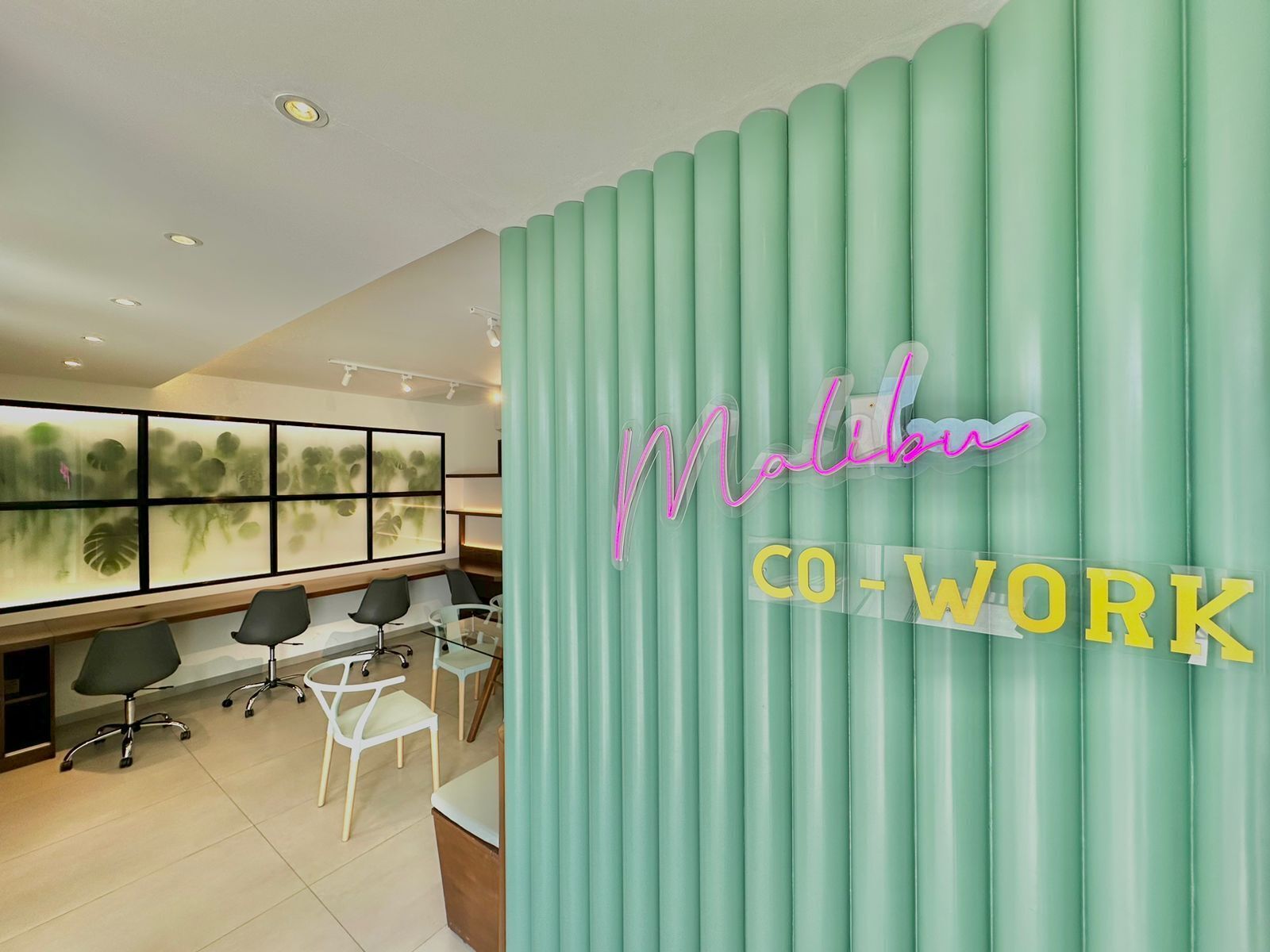 Un espacio de oficina moderno con una pared color turquesa, un letrero de neón que dice "Malibu CO-WORK" y un amplio ventanal que muestra follaje verde.