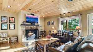 A cozy living room with a stone fireplace, wooden ceiling, and a large TV mounted above it.