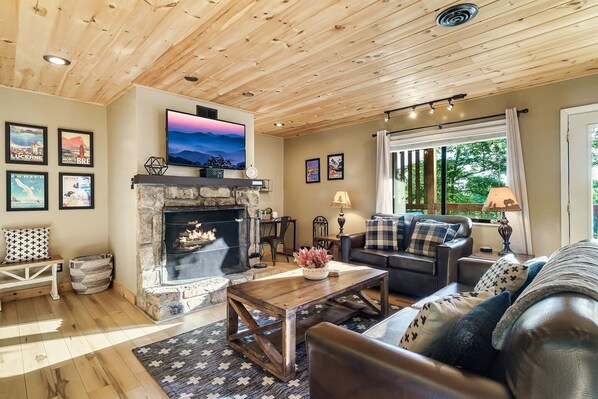 A cozy living room with a stone fireplace, wooden ceiling, and a large TV mounted above it.