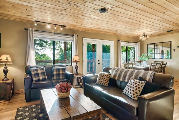 A cozy living room with a sofa, coffee table, and lamps. There is a dining area with a table and chairs visible through a glass door.