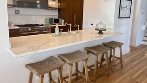 A kitchen with a marble island, wooden stools, and pendant lighting. - Luxury waterfront! The Pagoda Beach House (Oxnard)