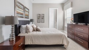 A bedroom with a bed, bedside table, lamp, dresser, TV, and artwork on the wall.
