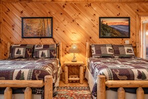 A cabin with two wooden beds, each with a patterned quilt and matching pillows, a small wooden nightstand with a lamp, and two framed pictures on the walls. - Free Dollywood Tix/Views/Pool/Spacious/Arcade (Sevierville)