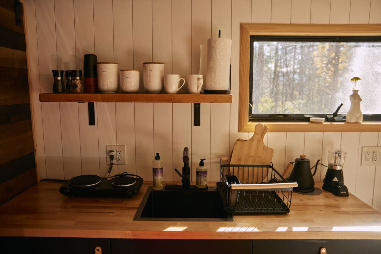 A kitchen with wooden shelves holding various kitchenware, a sink with a dish rack, and a window with a view of trees.