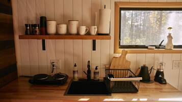 A kitchen with wooden shelves holding various kitchenware, a sink with a dish rack, and a window with a view of trees.