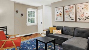 Una sala de estar con un sofá gris, un sillón rojo, una mesa de centro negra y una alfombra colorida. En la pared hay tres cuadros con motivos florales.