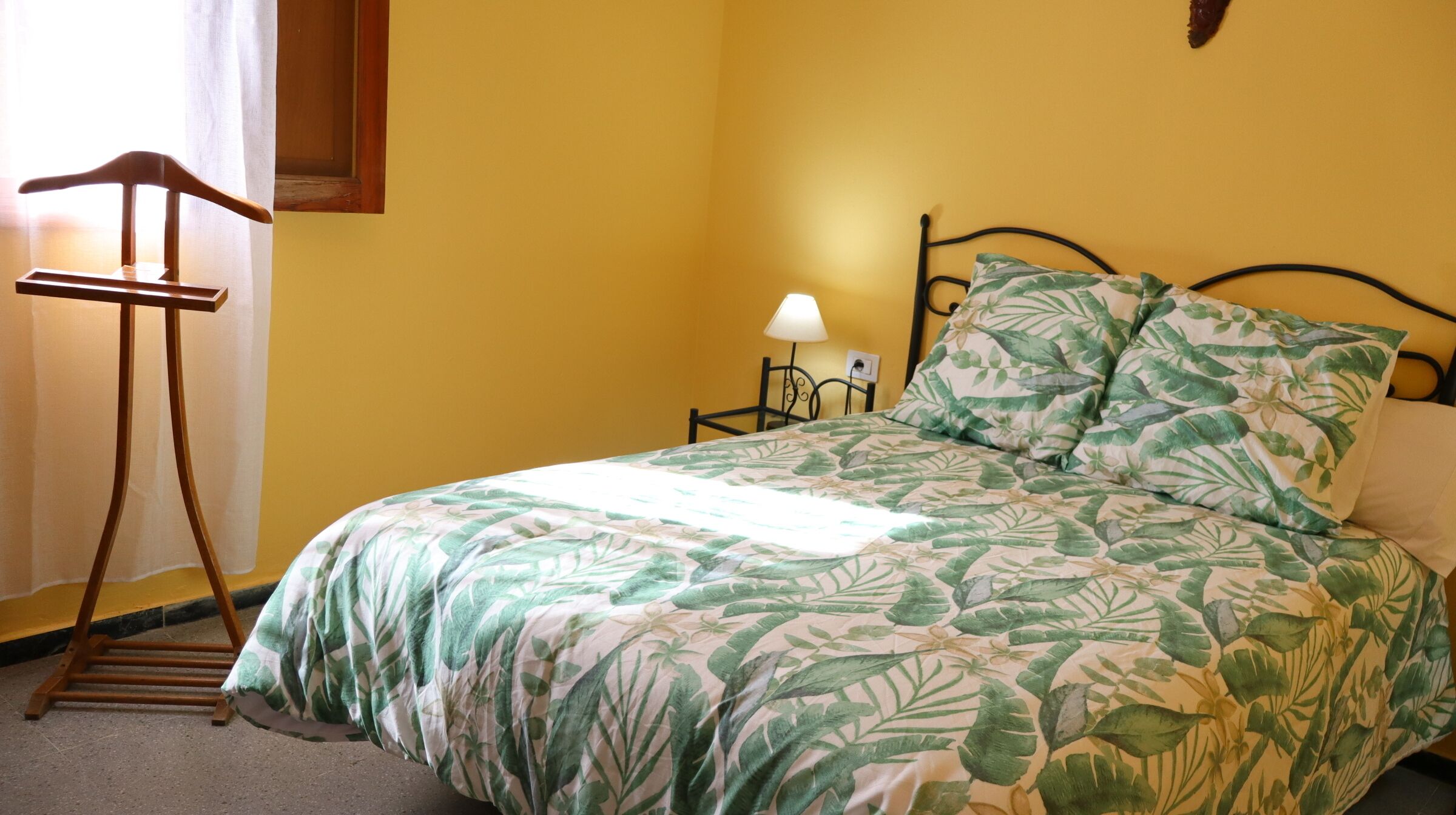 Ein Bett mit Bettdecke mit grünem Blattmuster, ein Holzhocker, ein Nachttisch mit Lampe und ein an der Wand befestigtes Regal.