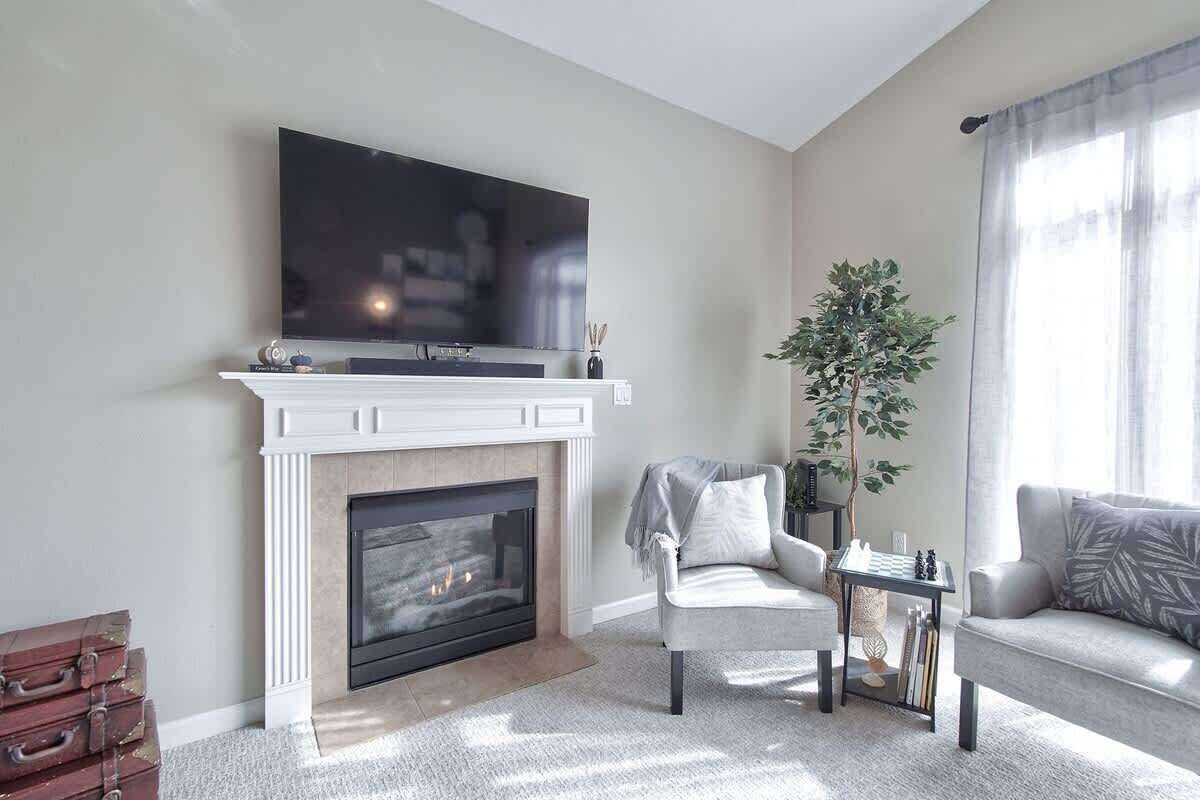 A living room with a fireplace, a flat-screen TV mounted above it, a grey sofa, a small side table with books, and a large potted plant.