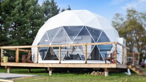 A geodesic dome structure with a wooden deck and glass panels, situated in a grassy area with trees in the background. - Skye's Dome - A relaxing getaway (Debert)