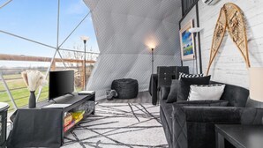 A modern living room with a large window, a black sofa, a TV, and a unique tent-like structure. - Skye's Dome - A relaxing getaway (Debert)