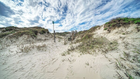 Uma praia com uma escadaria de madeira que leva a uma construção, cercada por dunas de areia e vegetação rasteira.