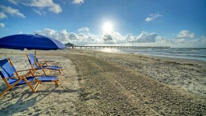 A beach with blue lounge chairs and a blue umbrella, a pier in the background, and the sun shining. - 2BR Condo w/ Gulf Views, 2 Pools, Courts Blue Water Bliss by Avantstay (Port Aransas)