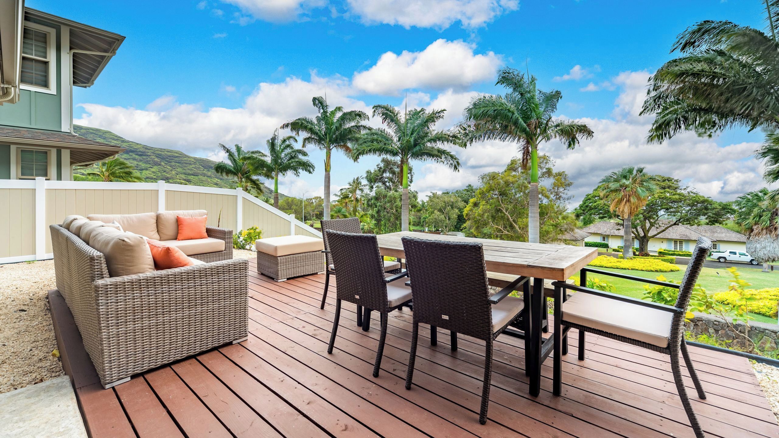 A deck with wicker furniture, a table, and chairs, overlooking a landscaped area with palm trees and a view of a house.