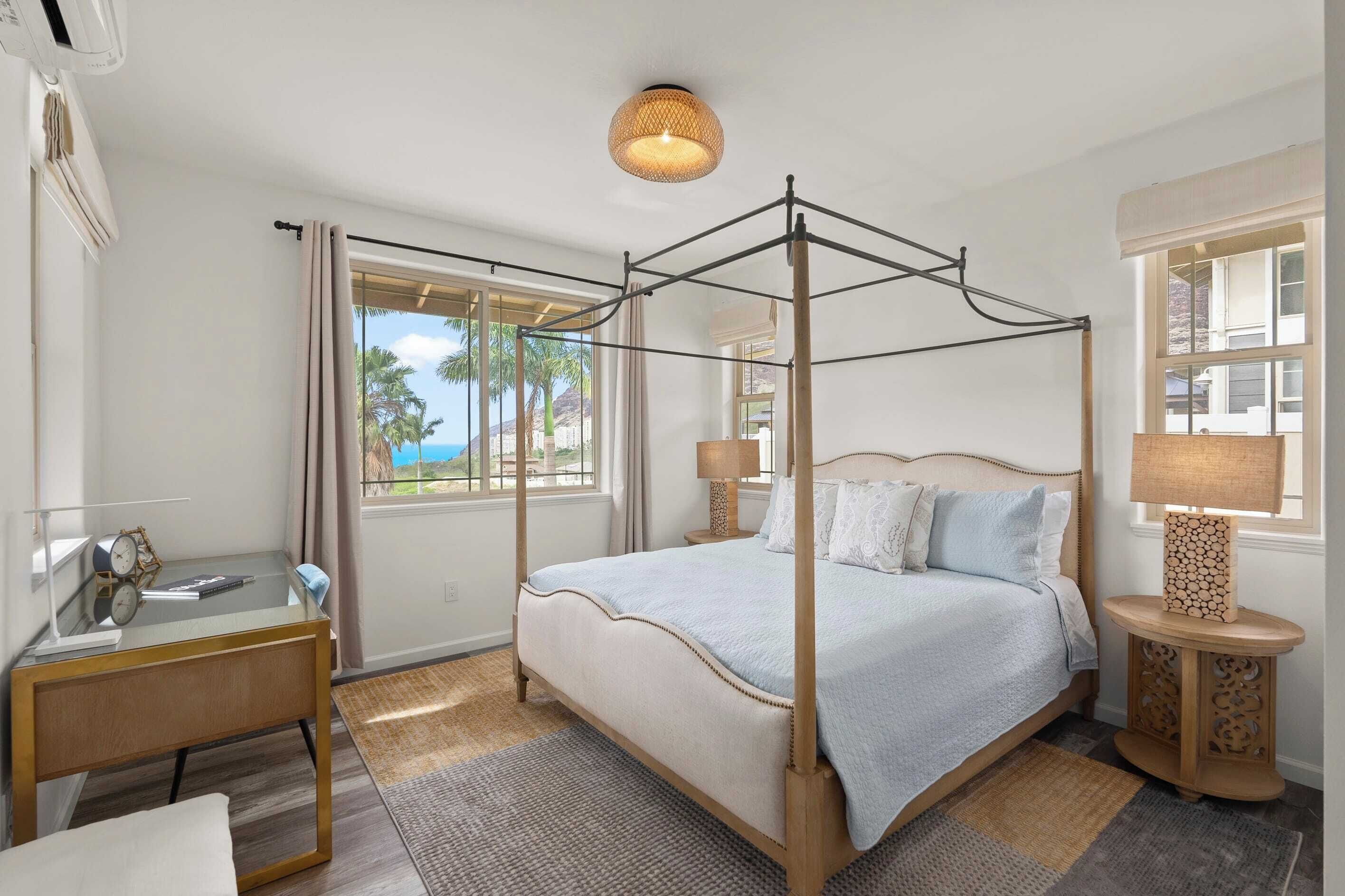 A four-poster bed with a view of palm trees, a desk with a lamp, and a window with curtains.
