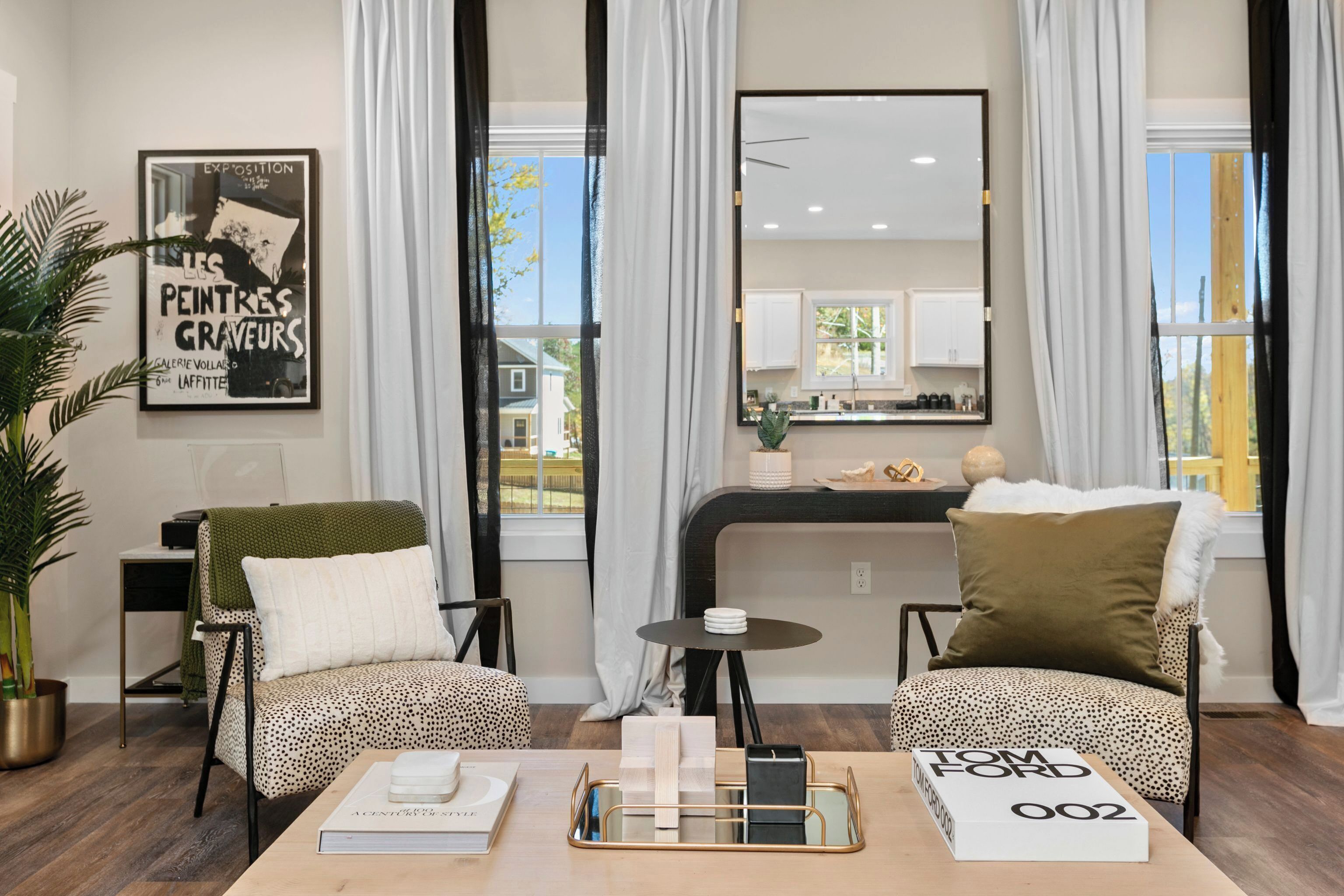 Ein modernes Wohnzimmer mit zwei Sesseln, einem Couchtisch mit Büchern und einer Serviertablett, einem gerahmten Kunstwerk an der Wand und Blick in die Küche durch ein großes Fenster.
