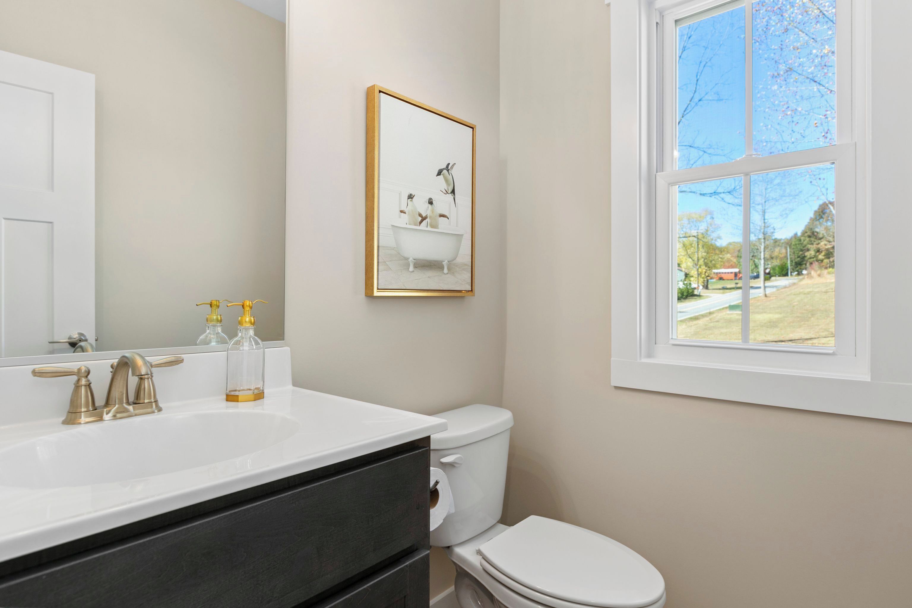 Une salle de bain avec un lavabo blanc, des robinets dorés, des toilettes et une fenêtre donnant sur des arbres et une maison.