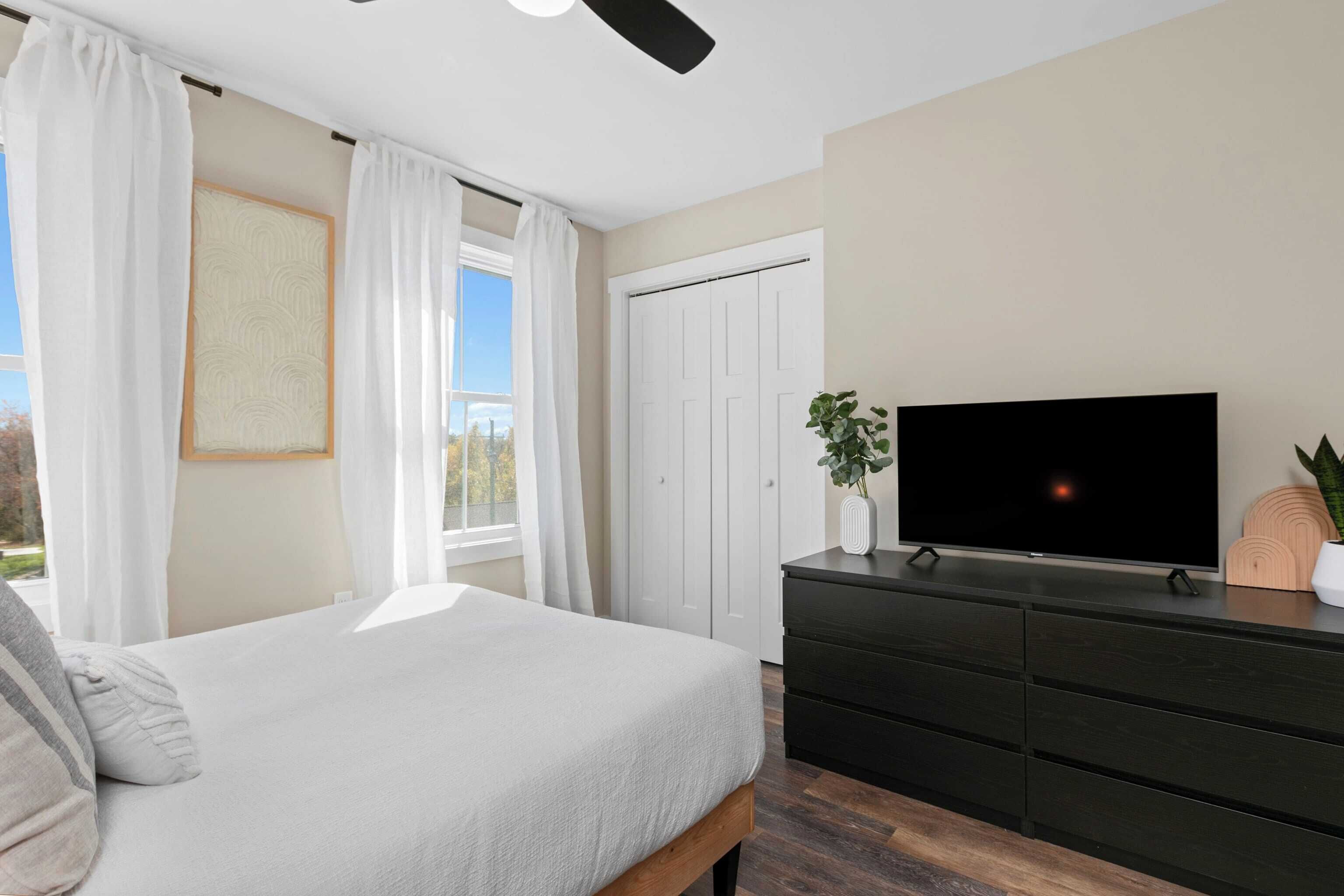 Une chambre à coucher avec un lit, une commode, un téléviseur et une fenêtre avec des rideaux.