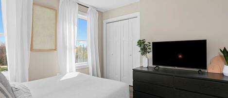 Une chambre à coucher avec un lit, une commode, un téléviseur et une fenêtre avec des rideaux.