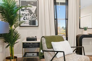 Ein Zimmer mit einer großen Kübelpflanze, einem Plattenspieler im Vintage-Stil, einem Punkte-Stuhl mit grüner Decke, einem Poster an der Wand und einem Fenster mit Vorhängen.