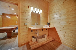 A wooden cabin bathroom with a wooden vanity, a mirror with lights, a sink, and a pool table in the adjacent room. - Java by Avantstay 12 Mins to Pigeon Forge + View (Sevierville)