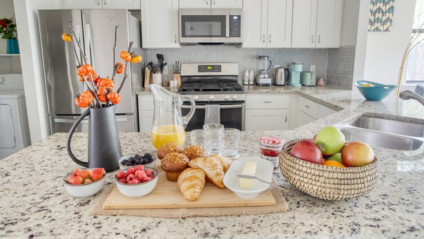 Una cocina con armarios blancos, microondas, estufa y diversos productos para el desayuno sobre la encimera.