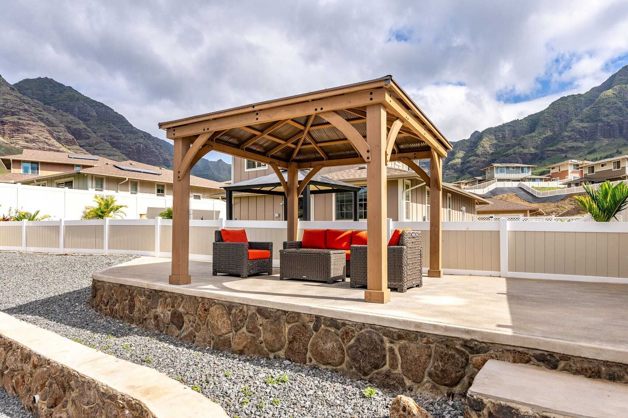 A covered outdoor seating area with wicker furniture and red cushions, set against a backdrop of mountains and residential buildings.