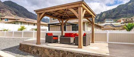 A covered outdoor seating area with wicker furniture and red cushions, set against a backdrop of mountains and residential buildings.