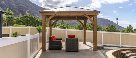 Uma área de estar coberta ao ar livre, com duas cadeiras de vime e uma pérgola de madeira, com vista para uma paisagem montanhosa.