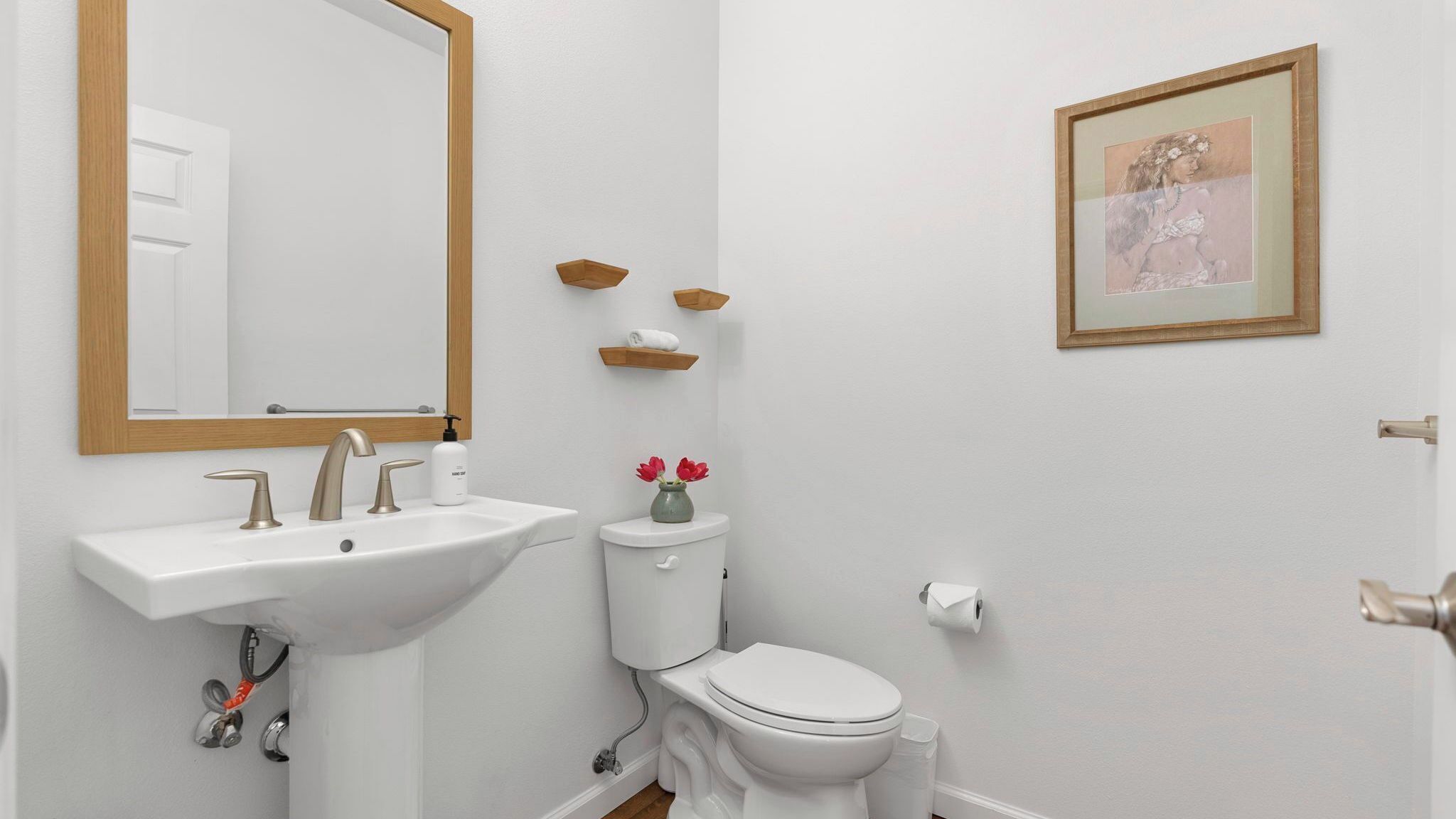 A bathroom with a white sink, a toilet, a mirror, a framed picture, and wooden shelves.