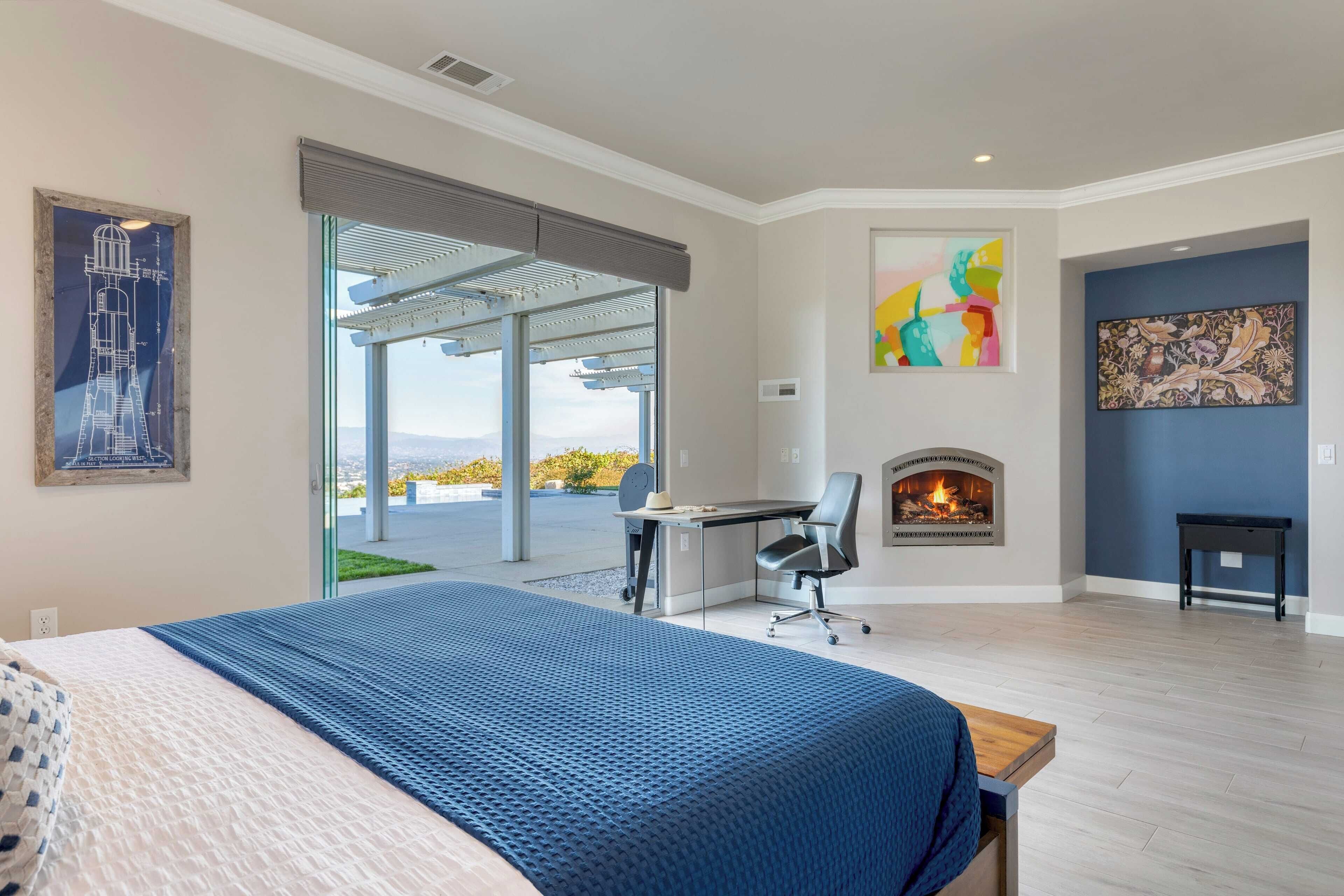 A modern bedroom with a large bed, a desk, a chair, and a fireplace.