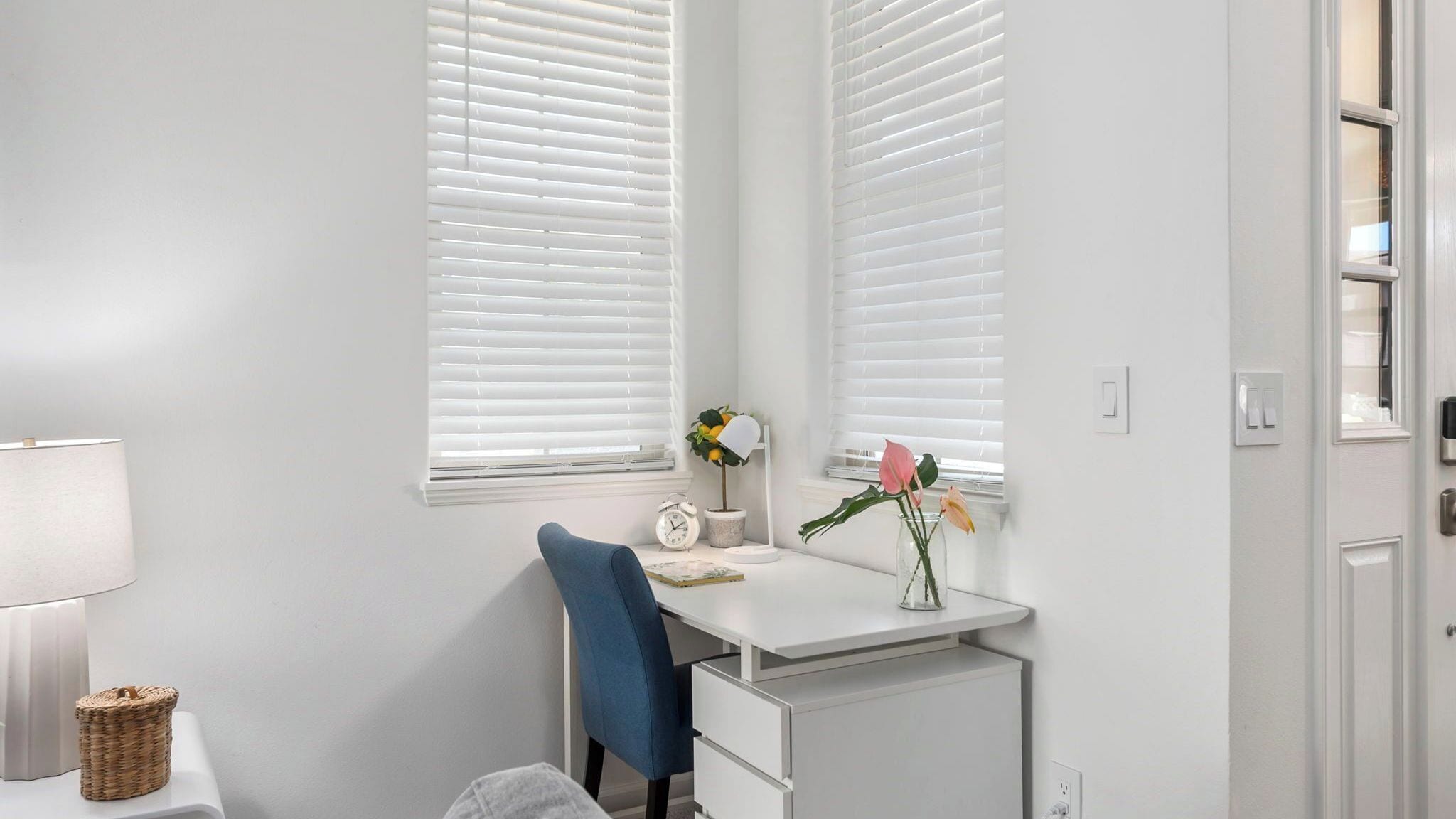 A white desk with a chair, a lamp, a clock, and a vase with flowers.